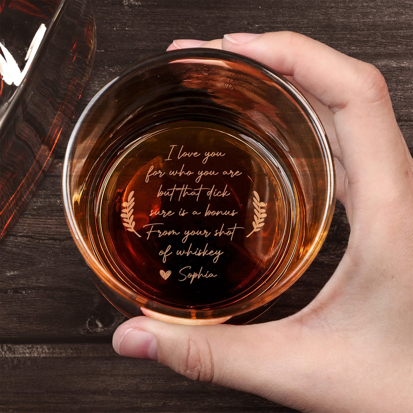 Personalized Whiskey Glass top view held in hand with amber whiskey and cheeky engraved love message at bottom on wooden table