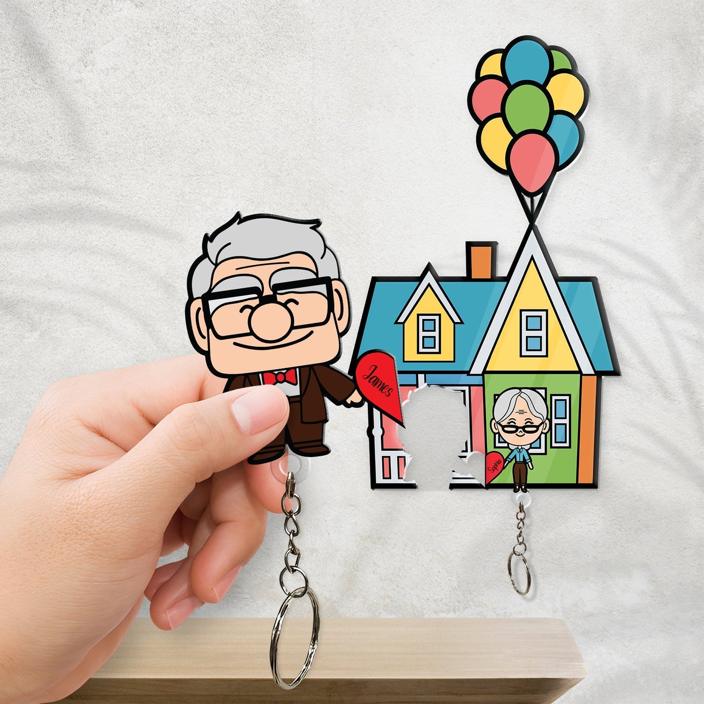 Hand holding a personalized couple keychain featuring cartoon elderly couple with colorful balloon house background