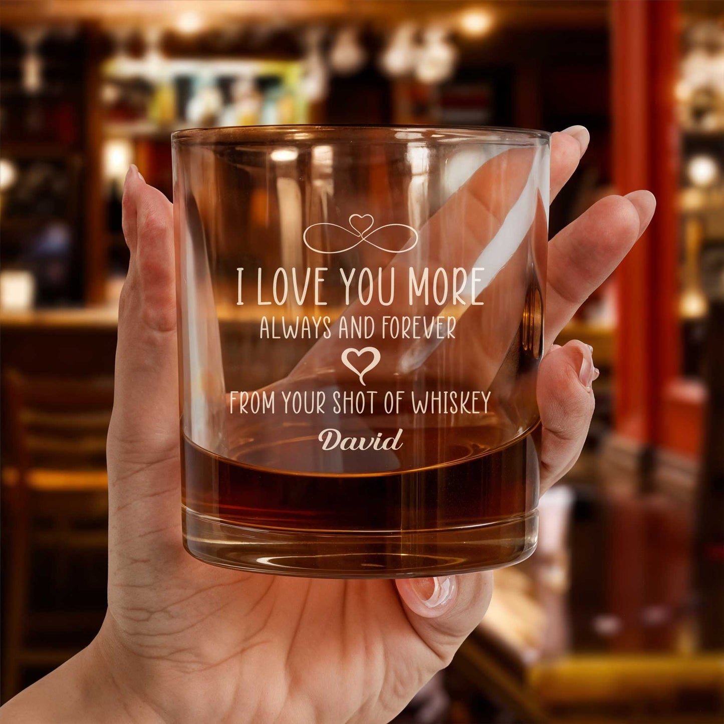 Personalized Whiskey Glass engraved I Love You More with name David held in hand with amber whiskey over bar backdrop