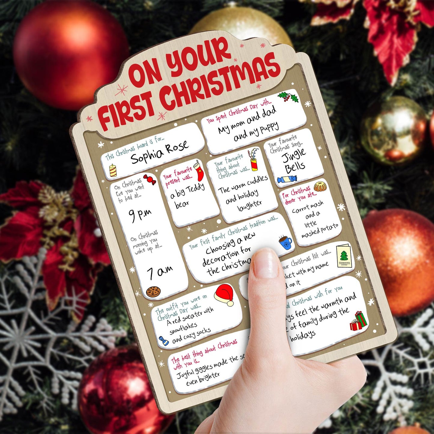 Personalized baby's first Christmas sign with festive notes and colorful holiday icons held against Christmas decorations